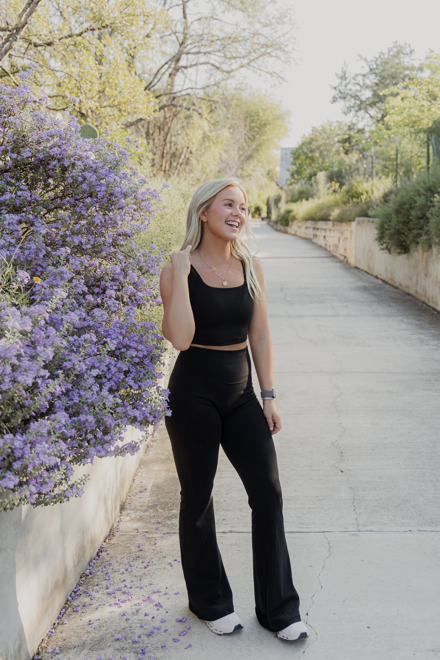 White women with blonde hair wearing a black tank top and flare leggings.  She is messing with her tank top while smiling at someone off the photo.