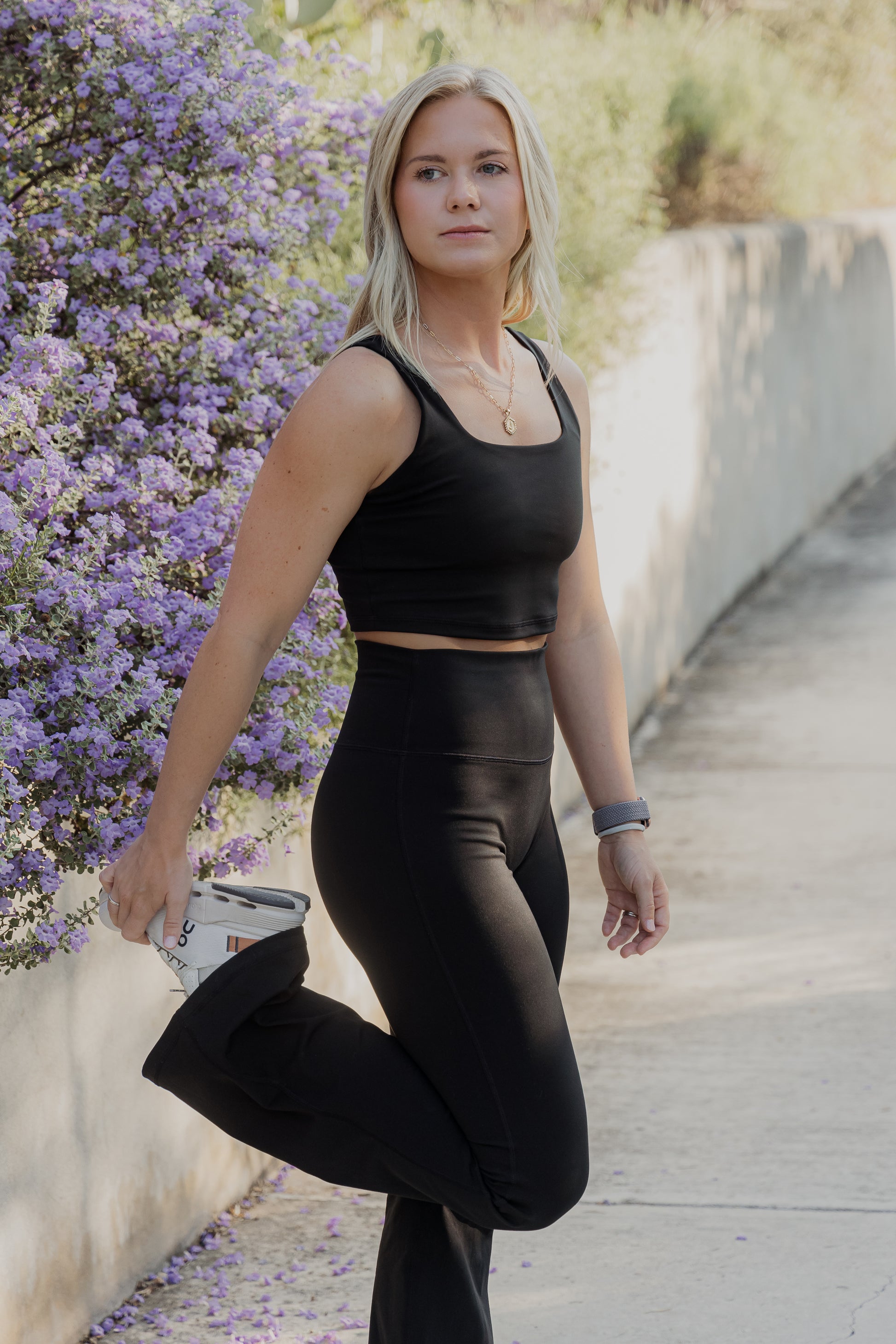 White women with blonde hair wearing a black tank top and flare leggings.  She is stretching her legs while looking off into the distance