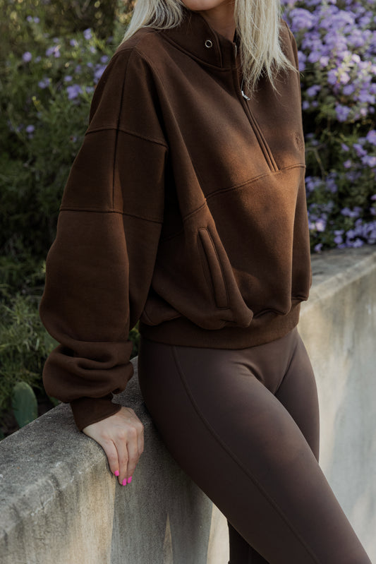 White women with blonde hair wearing a brown oversized cropped quarter zip sweater and brown leggings.  She is leaning on a concrete wall with purple flowers in the back ground.  It does not show her face but mainly the sweater.