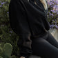 White women with blonde hair sitting  on a concrete wall with purple flowers in the back ground.  She is wearing a black oversized quarter zip sweater and black leggings.  This photo does not show her face or feet.