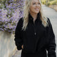 White women with blonde hair standing on a concrete sidewalk with purple flowers in the back ground.  She is wearing a black oversized quarter zip sweater and black leggings.  She is smiling but looking away from the camera.  