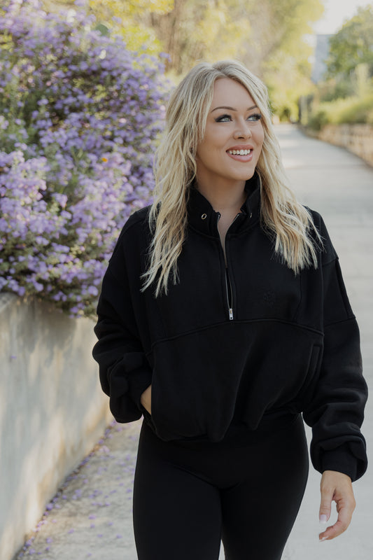 White women with blonde hair standing on a concrete sidewalk with purple flowers in the back ground.  She is wearing a black oversized quarter zip sweater and black leggings.  She is smiling but looking away from the camera.  