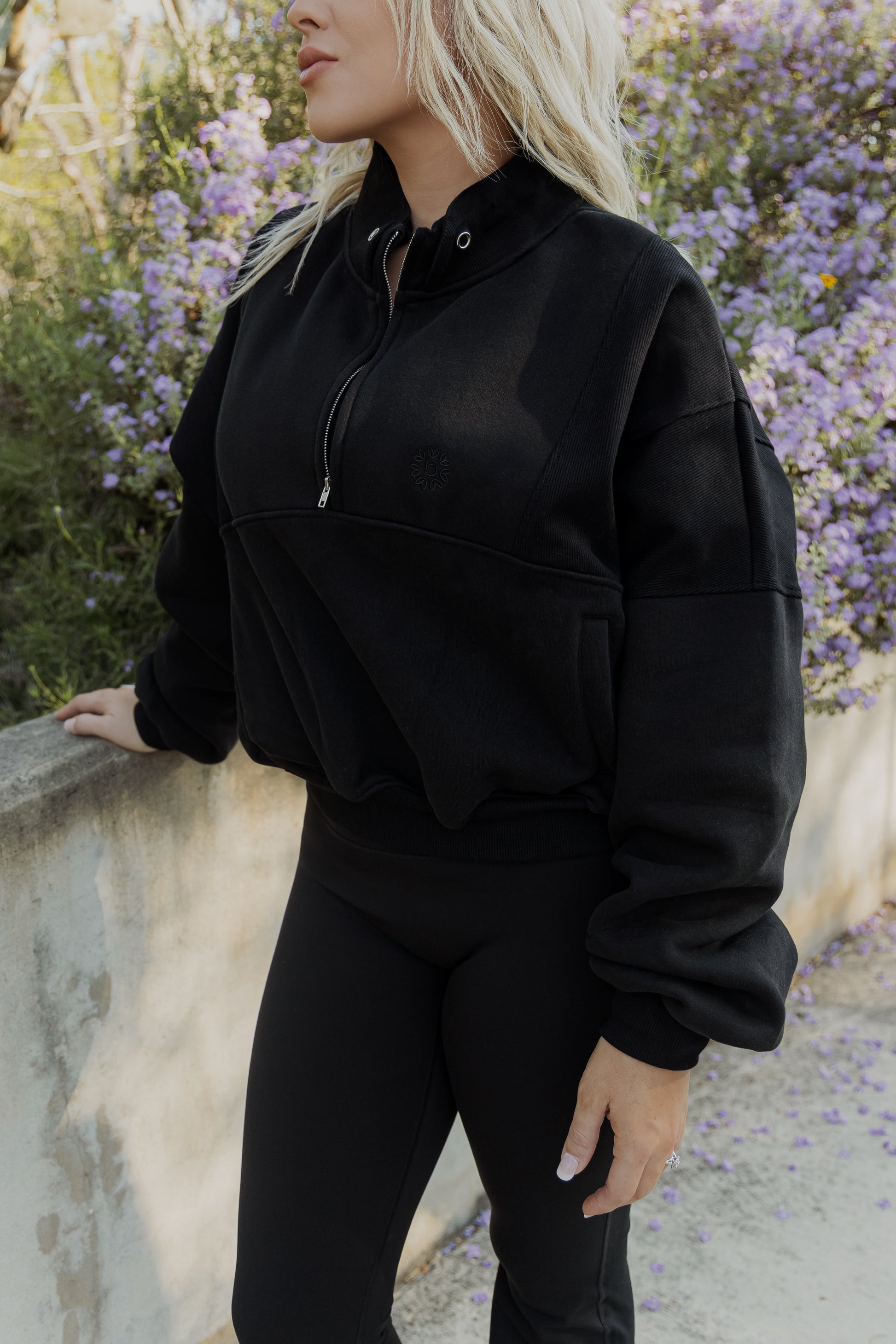 White women with blonde hair standing on a concrete sidewalk with purple flowers in the back ground.  She is wearing a black oversized quarter zip sweater and black leggings.  