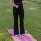 White women with blonde hair standing on a pink yoga mat that is laid in the grass.  she is wearing a black tank top sports bra and black flare leggings. 