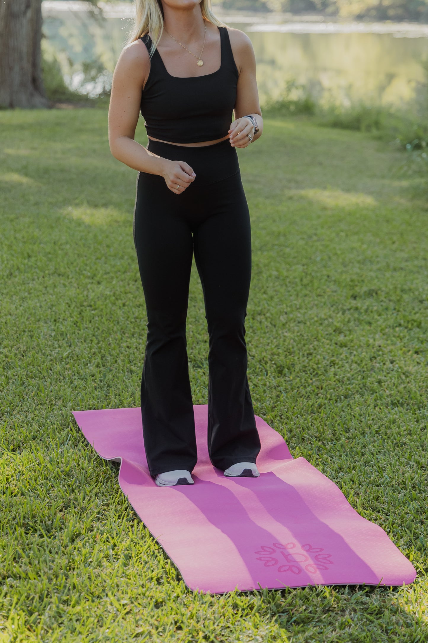 White women with blonde hair standing on a pink yoga mat that is laid in the grass.  she is wearing a black tank top sports bra and black flare leggings. 