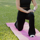 White women with blonde hair stretching her legs on a pink yoga mat that is laid in the grass.  She is wearing a black tank top sports bra and black flare leggings.