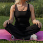 White women with blonde hair doing a yoga pose on a pink yoga mat that is laid in the grass.  She is wearing a black tank top sports bra, black flare leggings and white tennis shoes.