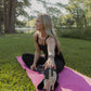 White women with blonde hair stretching her legs on a pink yoga mat that is laid in the grass.