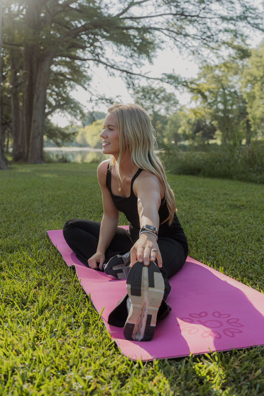 White women with blonde hair stretching her legs on a pink yoga mat that is laid in the grass.