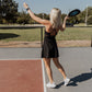 White women with blonde hair playing pickle ball.  she is holding the racket in her right arm and throwing the ball up with her left hand to show her serving the ball.  this photo is taken from the side/back.  She is wearing a black tennis dress and white tennis shoes while standing on a grey/red pickle ball court