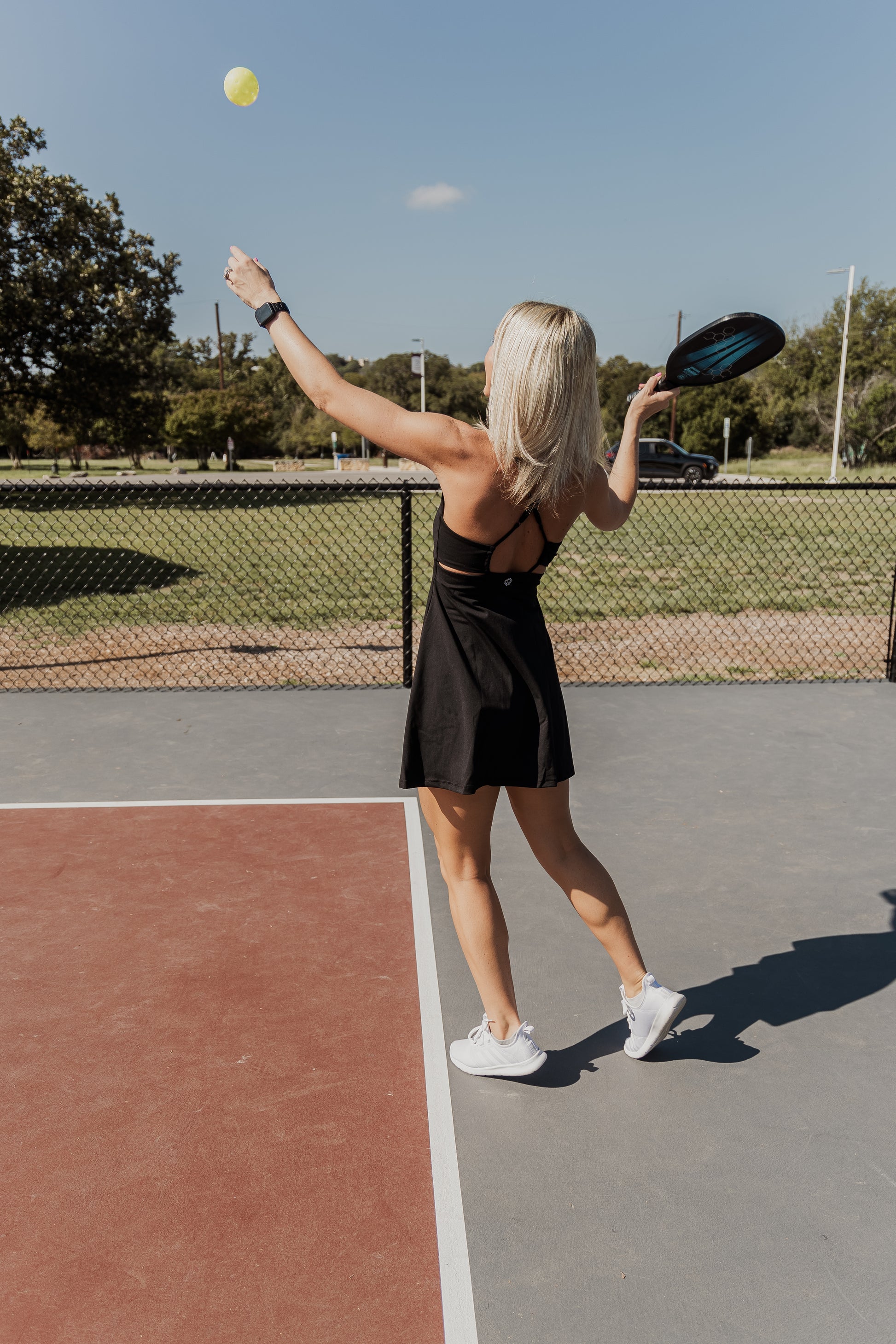 White women with blonde hair playing pickle ball.  she is holding the racket in her right arm and throwing the ball up with her left hand to show her serving the ball.  this photo is taken from the side/back.  She is wearing a black tennis dress and white tennis shoes while standing on a grey/red pickle ball court