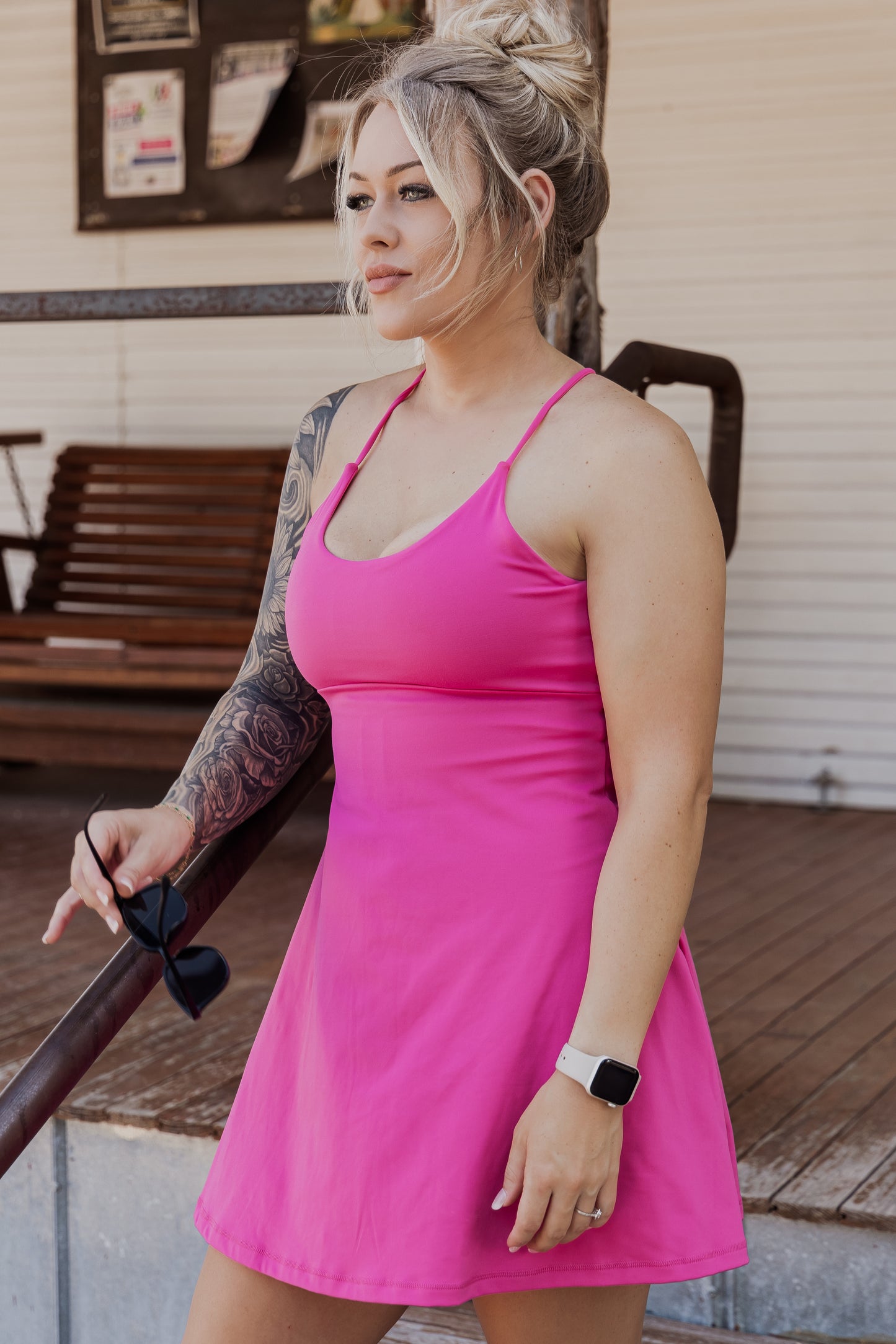 White women with blonde hair wearing it in a messy bun.  She is wearing a pink tennis dress and white van athletic shoes.  She is holding a pair of sunglasses in one hand.  The photo is taken from a side angle and she is not looking at the camera.  this photo does not show her legs just her upper body to the bottom of the dress.