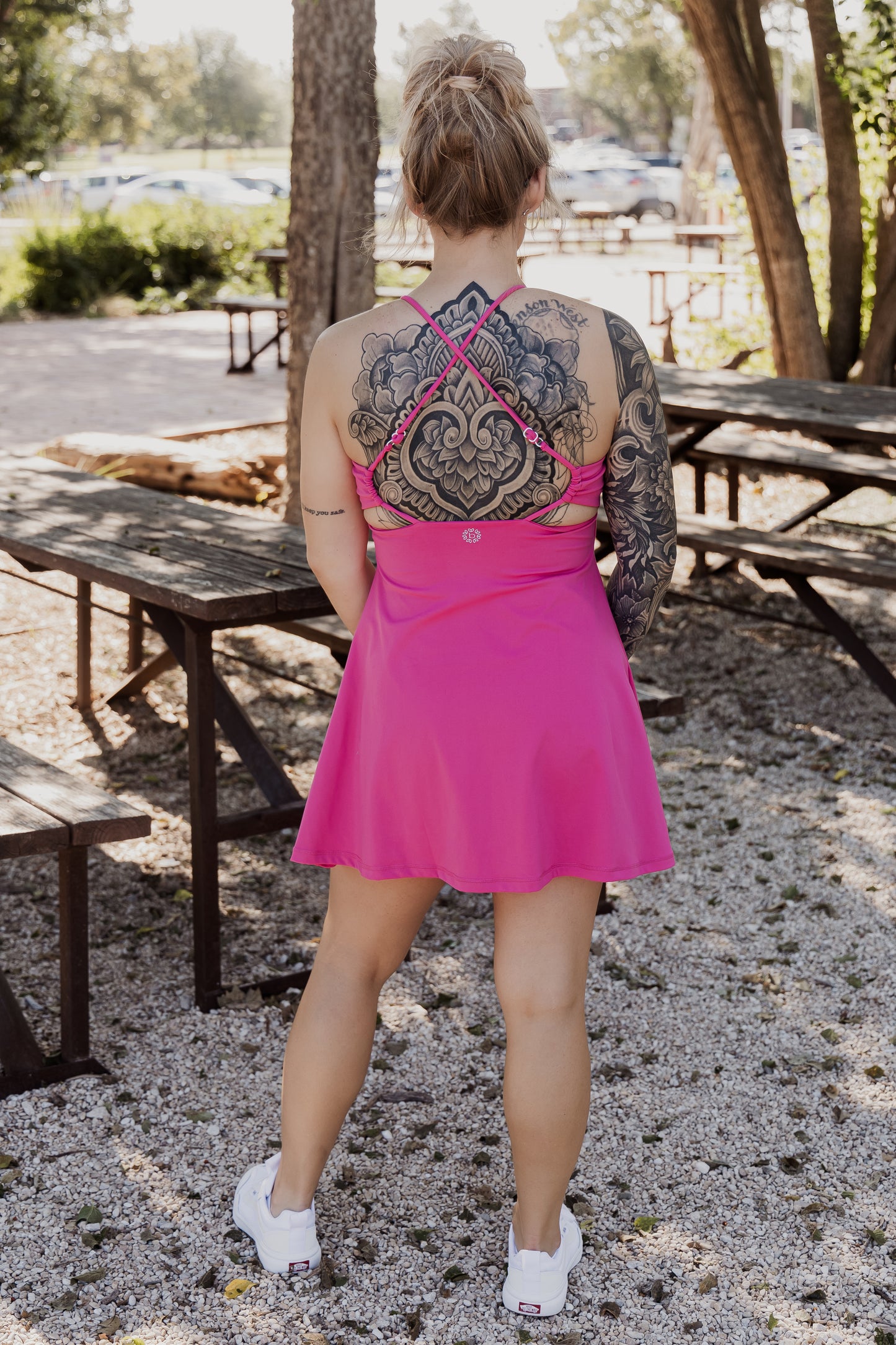 White women with blonde hair wearing it in a messy bun.  She is wearing a pink tennis dress and white van athletic shoes.  She is showing off the back of the dress.  She has visible tattoos on her back as well.