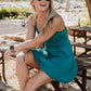 White women with blond curly hair sitting on top of a pick nik table with her arms rossed over one knee.  She is wearing a teal colored tennis dress and white and black checkered vans and sunglasses on her face.  she is looking off in the distance laughing