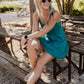 White women with blond curly hair sitting on top of a pick nik table with her arms rossed over one knee.  She is wearing a teal colored tennis dress and white and black checkered vans and sunglasses on her face.  she is looking off in the distance laughing