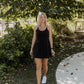 White women with blonde hair wearing a black tennis dress with white shoes.  She is walking towards the camera with her arms by her side and looking off into the distance with a smile on her face.