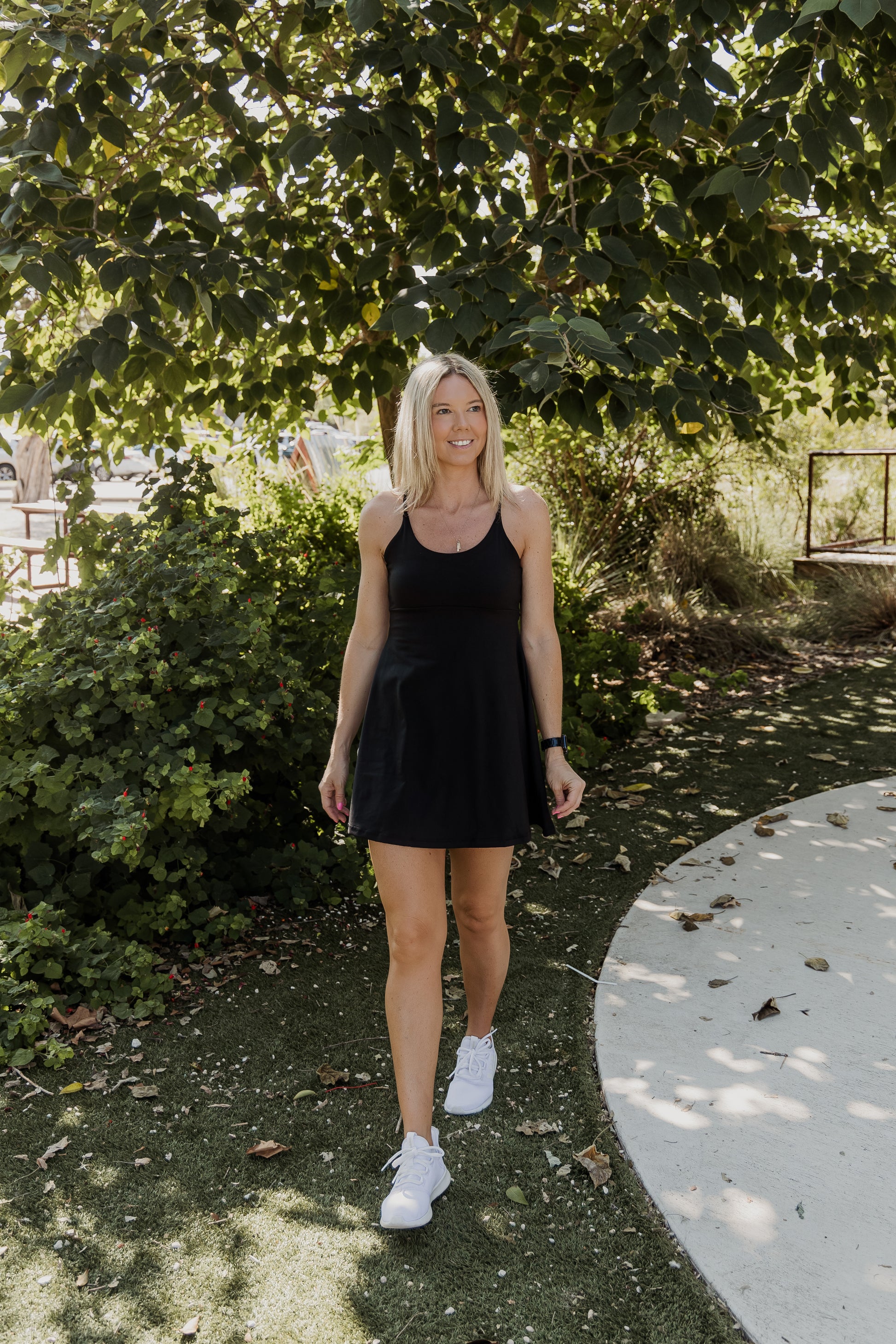 White women with blonde hair wearing a black tennis dress with white shoes.  She is walking towards the camera with her arms by her side and looking off into the distance with a smile on her face.