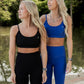 two white women with blonde hair standing next to the river at the park.  They are looking away from the camera and this photo only shows their upper half of their bodies.  they are both wearing matching legging and sports bra sets.  One is wearing an all black set and the other has on an electric blue set.  Both are shown wearing white tennis shoes.