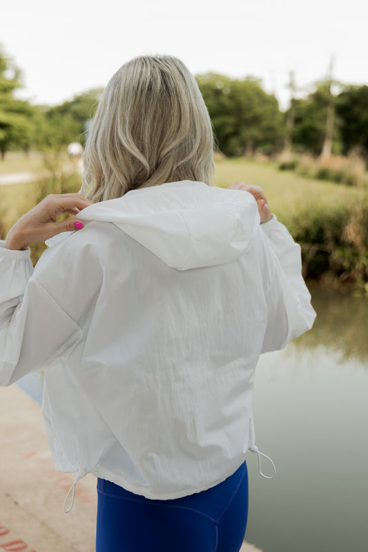White women with blonde hair wearing a white cropped hooded wind/rain jacket.  She is faced away from the camera and is showing off the hood on the jacket.