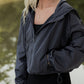 White women with blonde hair wearing a grey cropped hooded wind/rain jacket.  She is facing diagonal from the camera and is not showing her whole face.  Mainly its just an upclose shot of the jacket.