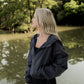 White women with blonde hair wearing a grey cropped hooded wind/rain jacket.  She is faced away from the camera and is standing infront of a river at the park.