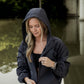 White women with blonde hair wearing a grey cropped hooded wind/rain jacket.  She has the hood pulled up over her head and is looking down zipping the jacket up.