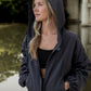 White women with blonde hair wearing a grey cropped hooded wind/rain jacket.  She has the hood over her head and is shown looking off into the distance.
