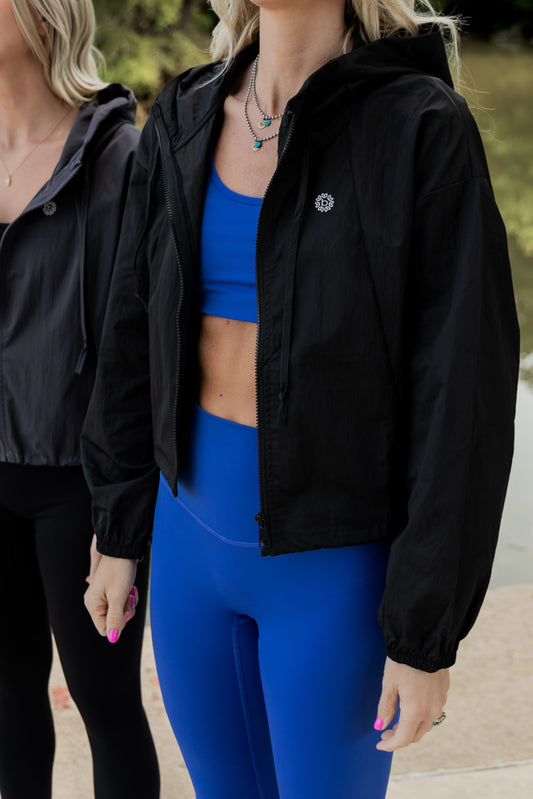 White women with blonde hair wearing a black cropped hooded wind/rain jacket.