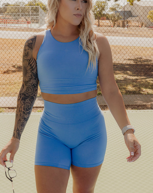 blonde haired women standing in the middle of a tennis court holding a pair of glasses in right hand.  she is wearing a matching bright blue workout set that includes a long lined tank top and biker shorts.