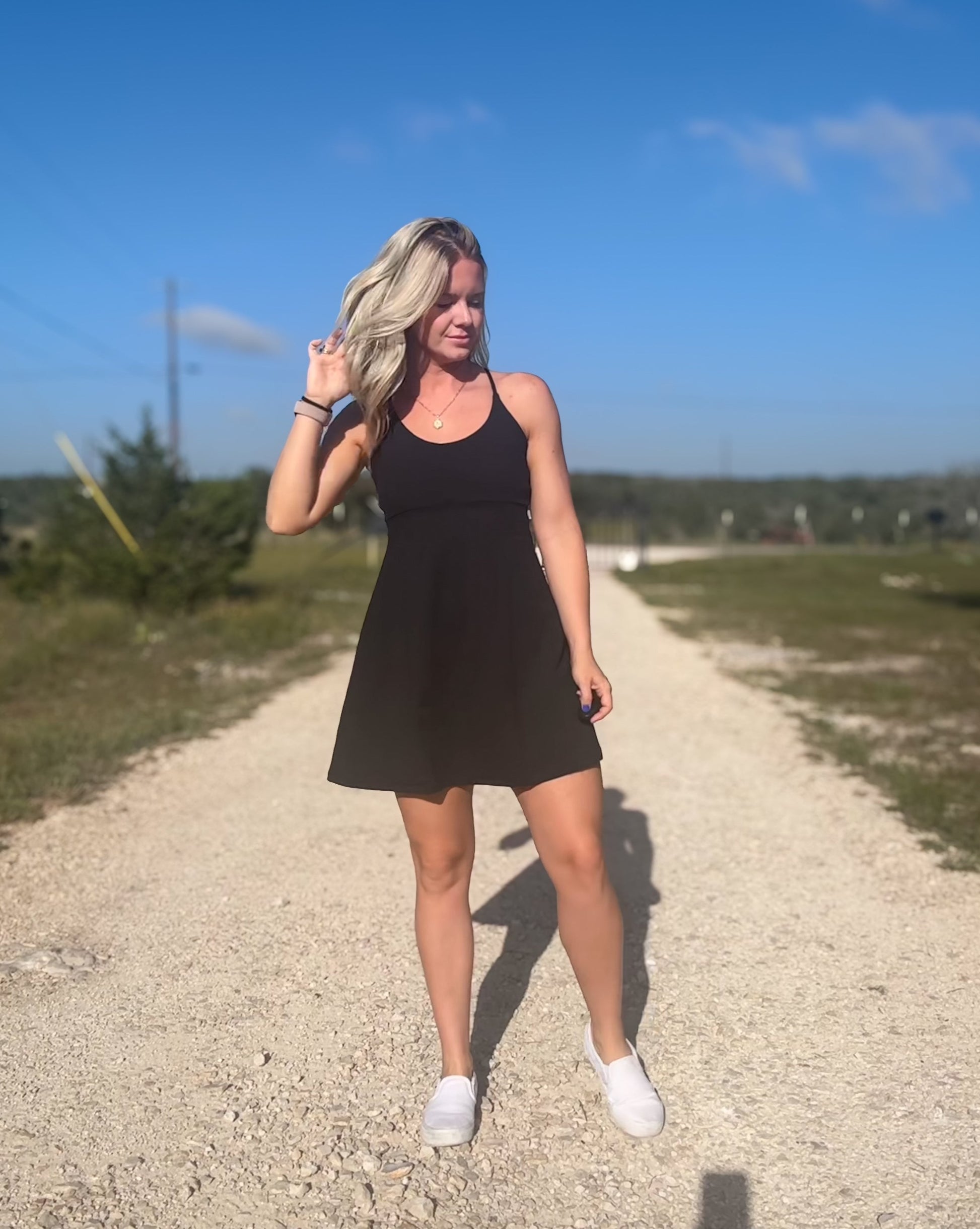 White blonde haired woman wearing a black activewear dress.  She is standing on a dirt road while wearing white vans