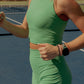Up close photo of a women running on a tennis court.  She is wearing a bright green activewear set that includes a long lined tank top and biker shorts.