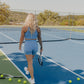 Blonde curly haired women walking away from the camera while standing on a tennis court holding a tennis racket in her right hand.  She is wearing a long lined tank top and biker shorts in the color bright blue.