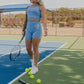 blonde haired women standing in the middle of a tennis court.  she is holding a tennis racket in her right hand.  She is wearing a matching workout set that includes a long lined tank top and matching biker shorts in the bright blue color.