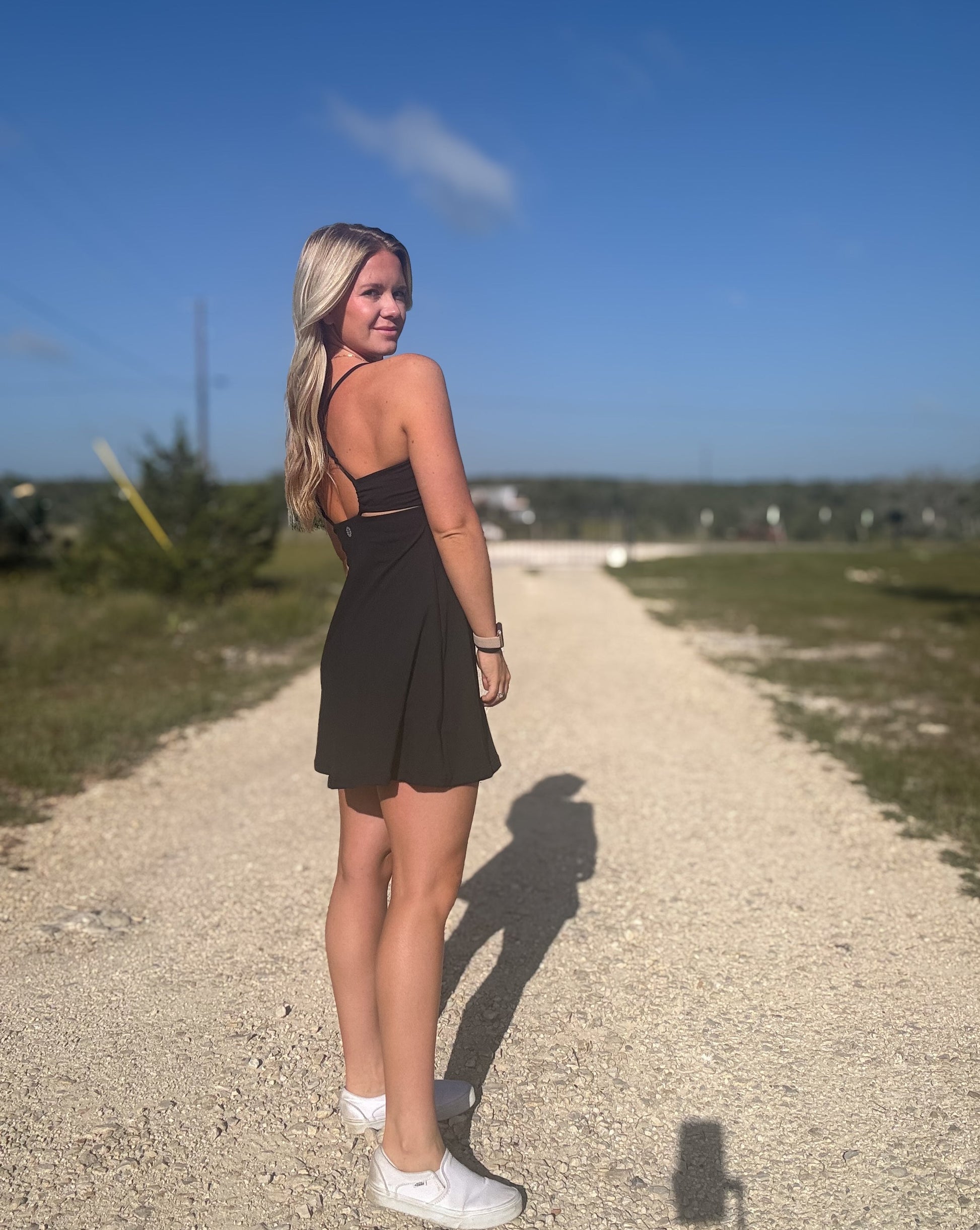 White blonde haired woman wearing a black activewear dress.  She is standing on a dirt road while wearing white vans