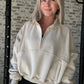 white women wearing an oversized half zip pull over in a tan color.  This women is standing in front of a white washed brick wall.