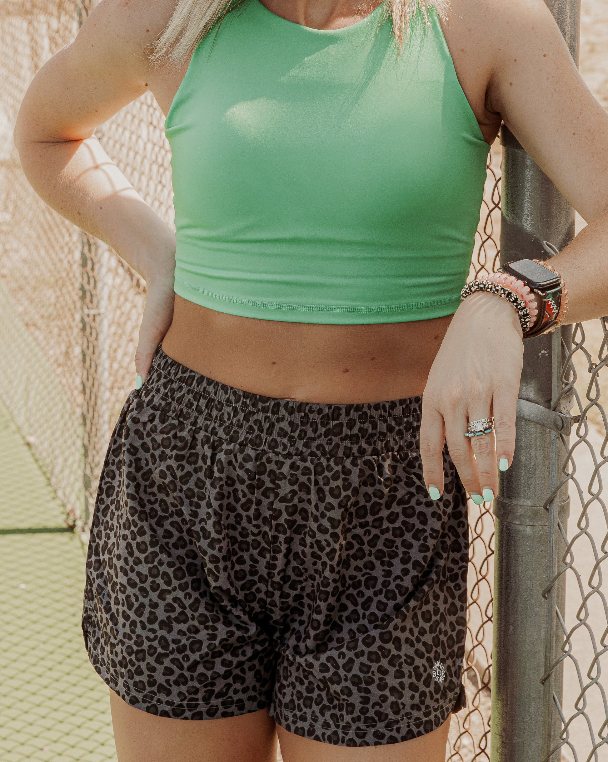 Blonde haired women standing on a tennis court next to the fence. She is wearing a bright green long lined tank top and high waisted grey leopard running shorts. This photo does not include her face but the middle half of her body.