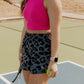 blonde haired women standing in the middle of a tennis court.  she is holding a tennis racket in one hand and a tennis ball in the other.  She is wearing a bright pink long lined tank top and a pair of printed running shorts in a panther printing.  These are high waisted and have a built in panty liner.