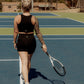 Blonde haired women walking on a tennis court with a tennis racket in her hand.  She is looking away from the camera the camera and has her hair up.  She is wearing a matching tank top and biker short set in the color black.