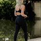 white women with blonde hair standing next to the river at the park.  she is facing away from the camera and is wearing a matching legging and sports bra set.  the leggings have a deep v in the stitching on the bottom and the sports bra has a twist back and adjustable straps.  she is wearing white tennis shoes and is shown walking.