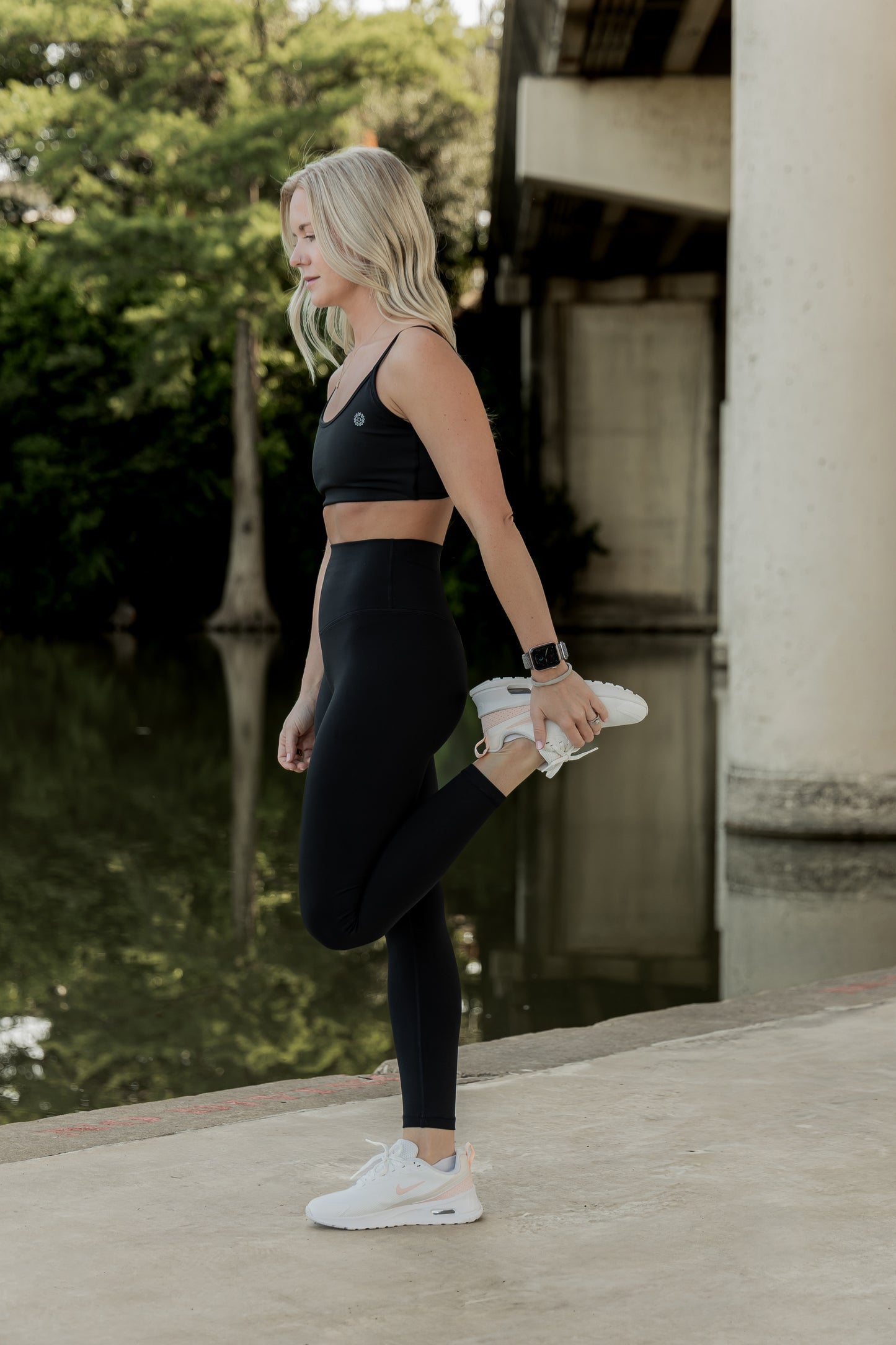 white women with blonde hair stretching her legs by the river at the park.  she is wearing a matching legging and sports bra set in black.  she is wearing white nike tennis shoes.