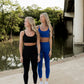 two white women with blonde hair standing next to the river at the park.  they are both wearing matching legging and sports bra sets.  One is wearing an all black set and the other has on an electric blue set.  Both are shown wearing white tennis shoes.